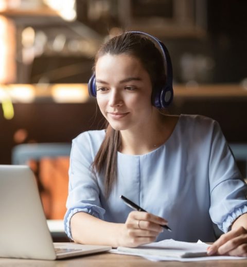 Focused woman wearing headphones using laptop in cafe, writing notes, attractive female student learning language, watching online webinar, listening audio course, e-learning education concept